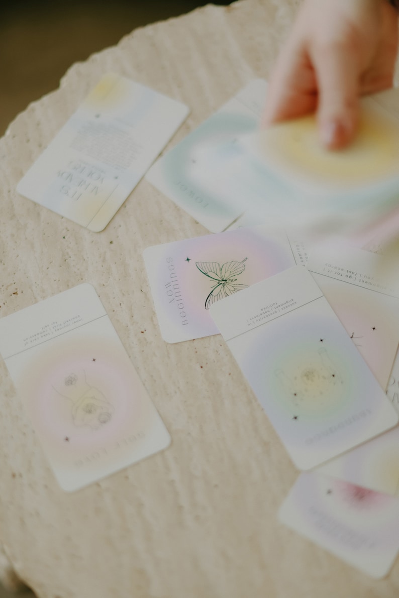 May include: A set of pastel-colored oracle cards with illustrations of butterflies, stars, and other celestial imagery. The cards have text on them, including the words "Realignments" and "Reconnecting".