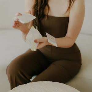 May include: A person is holding a deck of cards with a light blue, yellow, and white design. Three cards are laid out on a light beige table. The cards have a light blue, light blue, and pink design.