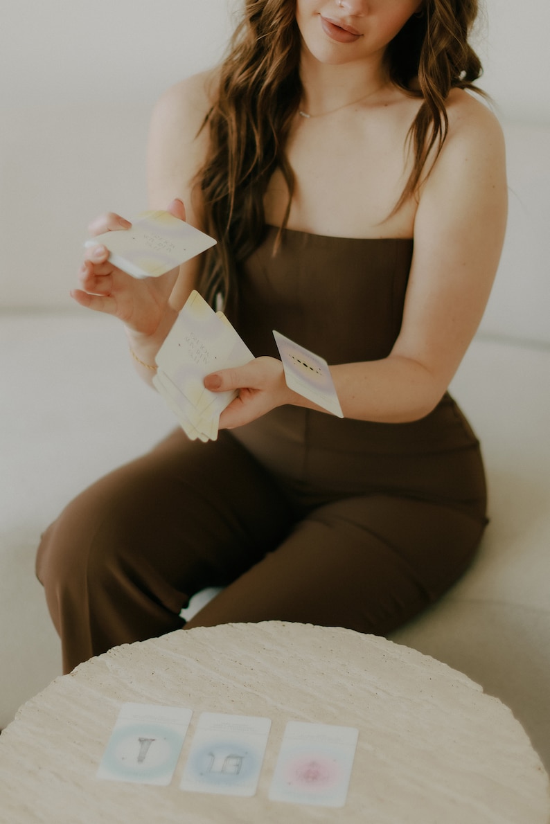 May include: A person is holding a deck of cards with a light blue, yellow, and white design. Three cards are laid out on a light beige table. The cards have a light blue, light blue, and pink design.