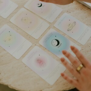 May include: A set of colorful oracle cards with illustrations of celestial bodies, a swan, and a crystal ball. The cards have a gradient background and are laid out on a light beige surface.