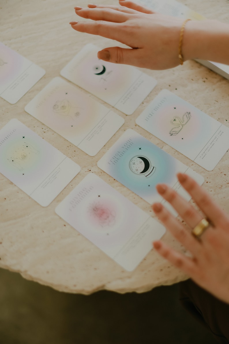 May include: A set of colorful oracle cards with illustrations of celestial bodies, a swan, and a crystal ball. The cards have a gradient background and are laid out on a light beige surface.