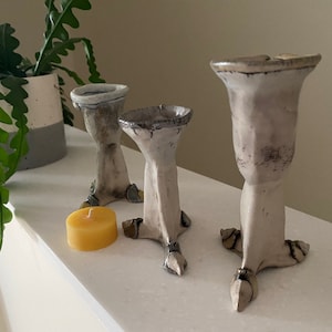 Peut inclure: Trois bougeoirs en céramique avec un glaçage blanc et une finition texturée. Les bougeoirs sont en forme de pied d'oiseau à trois doigts. Une petite bougie jaune se trouve devant les bougeoirs.