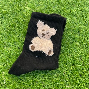 May include: Black socks with a white teddy bear design. The bear is embroidered on the socks and has a brown nose and eyes.