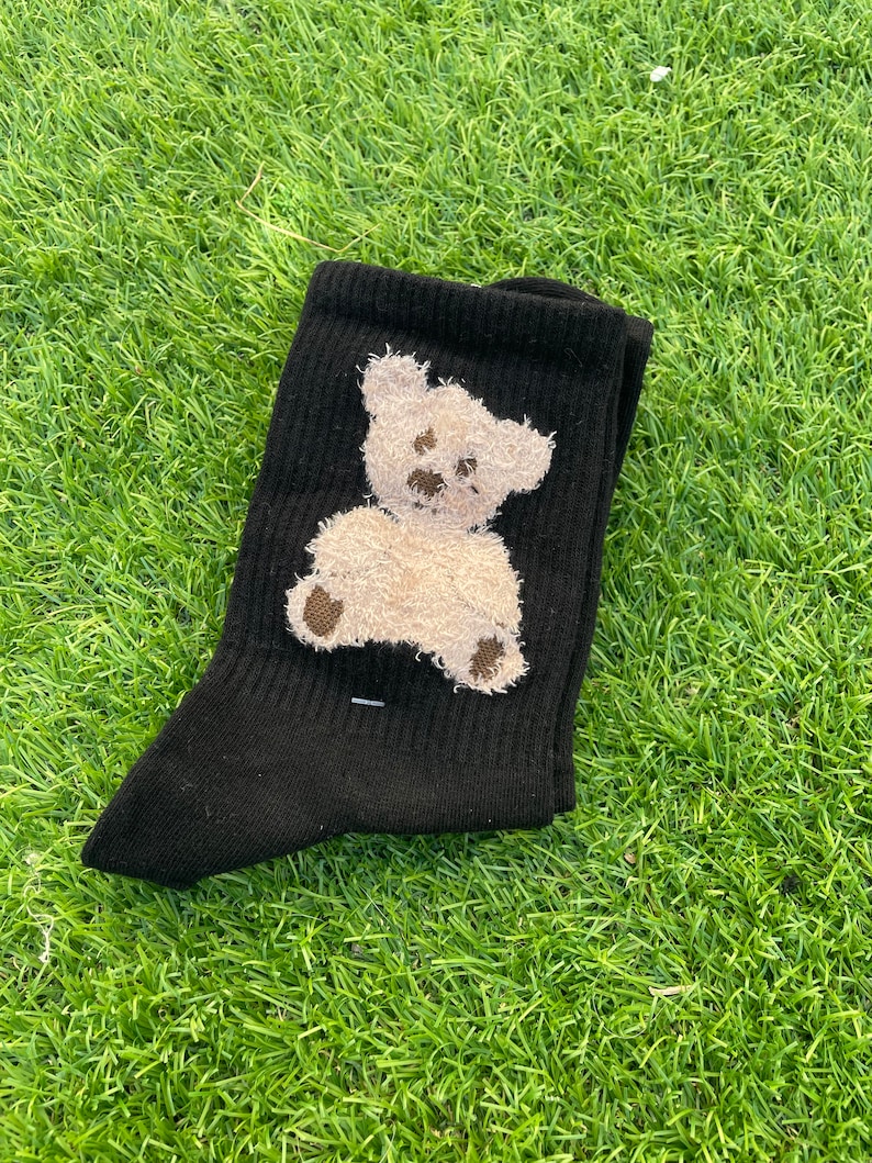May include: Black socks with a white teddy bear design. The bear is embroidered on the socks and has a brown nose and eyes.