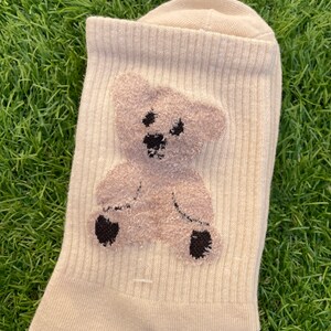 May include: A pair of beige socks with a brown teddy bear design. The bear is fuzzy and has black eyes and a black nose.