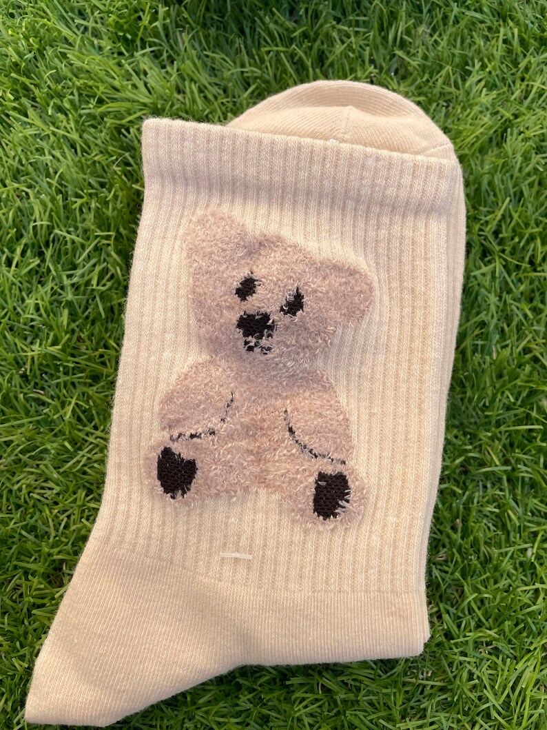 May include: A pair of beige socks with a brown teddy bear design. The bear is fuzzy and has black eyes and a black nose.