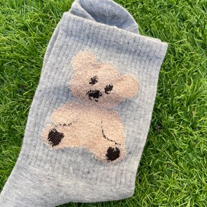 May include: A pair of gray socks with a brown teddy bear design. The teddy bear is embroidered on the socks and has a fuzzy texture.
