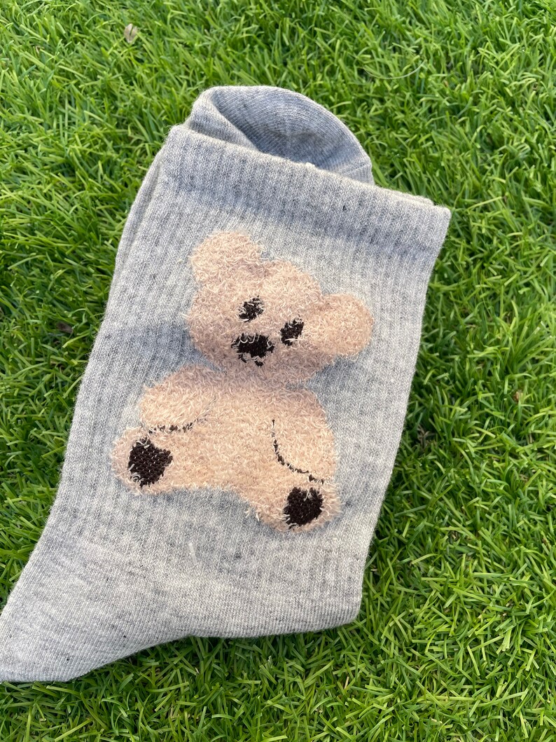May include: A pair of gray socks with a brown teddy bear design. The teddy bear is embroidered on the socks and has a fuzzy texture.
