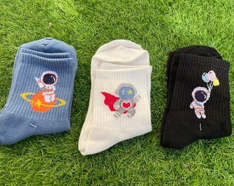 May include: Three pairs of socks with astronaut designs. One pair is blue with an astronaut in a space suit holding a planet. One pair is white with an astronaut in a space suit wearing a red cape. One pair is black with an astronaut in a space suit holding balloons.