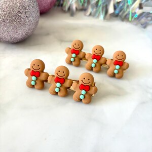 May include: Five gingerbread man hair clips with a gold bar connecting them. The gingerbread men are brown with white icing and red bows. The gingerbread men have blue eyes and a smiling face.