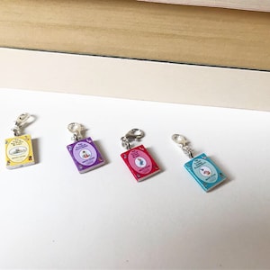 Op de afbeelding: Vier boekvormige bedels met zilveren sluitingen. De bedels zijn versierd met afbeeldingen van kinderboeken, waaronder "Het verhaal van Peter Konijn" en "De Very Hungry Caterpillar".