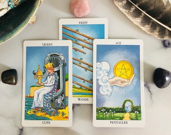 May include: A tarot card spread featuring the Queen of Cups, Eight of Wands, and Ace of Pentacles. The cards are surrounded by crystals, a feather, and a shell with sage. The cards have a white border and feature colorful illustrations.