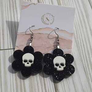 Puede incluir: Un par de pendientes de flores negras con purpurina y una calavera blanca en el centro de cada flor. Los pendientes tienen ganchos plateados. El fondo incluye una tarjeta con el texto "stars and serenity".