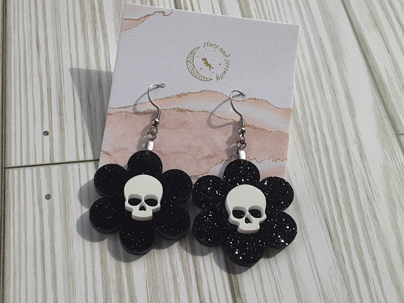 Puede incluir: Un par de pendientes de flores negras con purpurina y una calavera blanca en el centro de cada flor. Los pendientes tienen ganchos plateados. El fondo incluye una tarjeta con el texto "stars and serenity".