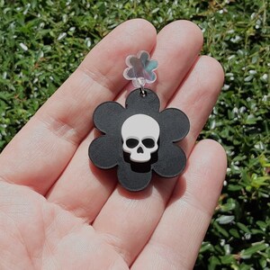Puede incluir: Un pendiente negro en forma de flor con una calavera blanca en el centro. El pendiente se sostiene en una mano sobre un fondo de follaje verde. El pendiente tiene una parte superior transparente en forma de flor iridiscente.