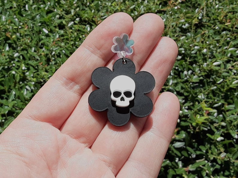 Puede incluir: Un pendiente negro en forma de flor con una calavera blanca en el centro. El pendiente se sostiene en una mano sobre un fondo de follaje verde. El pendiente tiene una parte superior transparente en forma de flor iridiscente.