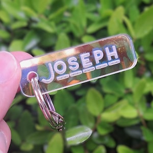 May include: A clear rectangular keychain with the name "JOSEPH" in white and blue lettering. The keychain is attached to a silver key ring. The background is blurred green foliage.