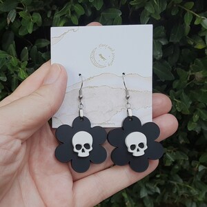 Puede incluir: Pendientes negros con forma de flor, con una calavera blanca en el centro. Los pendientes tienen ganchos plateados y se exhiben en una tarjeta blanca con texto dorado que dice "stars and serenity".