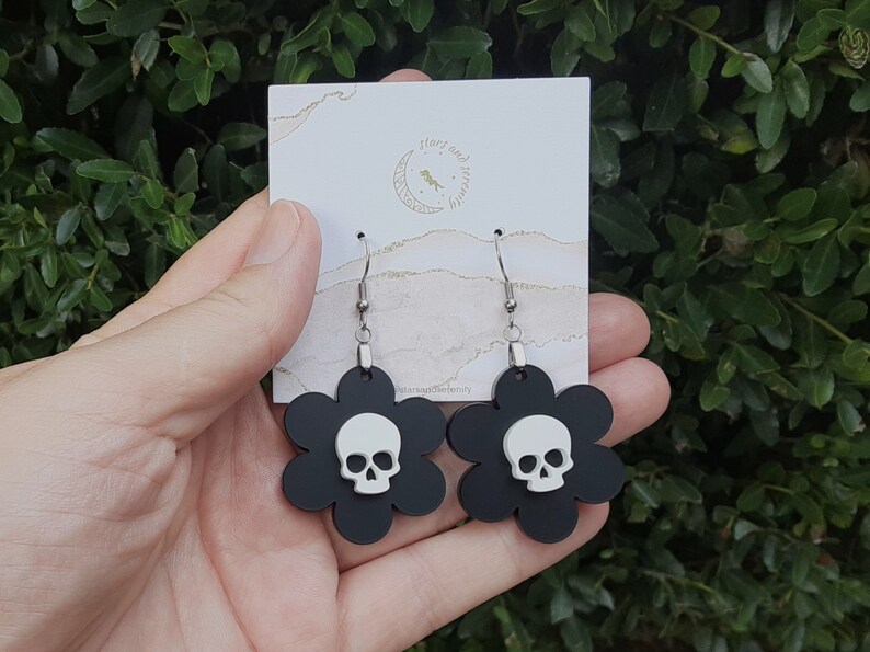 Puede incluir: Pendientes negros con forma de flor, con una calavera blanca en el centro. Los pendientes tienen ganchos plateados y se exhiben en una tarjeta blanca con texto dorado que dice "stars and serenity".