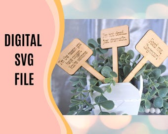 May include: Three wooden plant markers with funny sayings. The markers are brown and have white text. The text on the markers reads: "I'm the reason you have oxygen. You're welcome", "I'm not dead... Just dramatic." and "Feed me, Seymour!"