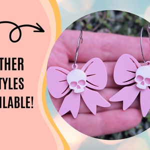 Puede incluir: Pendientes de lazo rosa con un dije de calavera blanca. Los pendientes cuelgan de aros plateados. La imagen tambi&eacute;n incluye el texto "OTHER STYLES AVAILABLE!" en letras negras.