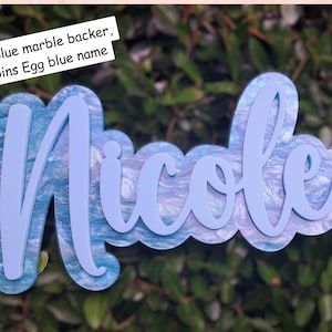 Puede incluir: Un recorte de nombre en capas con un fondo de mármol azul cielo y un nombre azul huevo de petirrojo. El nombre "Nicole" está en una fuente cursiva. El recorte se encuentra sobre un fondo de follaje verde.