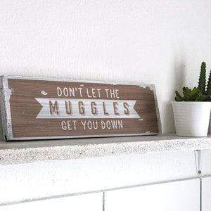 Könnte beinhalten: Ein rustikales Holzschild mit dem Text "Don't let the Muggles get you down". Das Schild ist braun mit weißen Buchstaben und steht auf einem weißen Regal mit einer kleinen Sukkulente im Topf rechts.