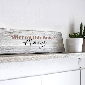 Könnte beinhalten: Ein weiß gestrichenes Holzschild mit dem Text "After all this time? Always" in rosa und schwarzen Buchstaben. Das Schild steht auf einem weißen Regal mit zwei kleinen Topfpflanzen.