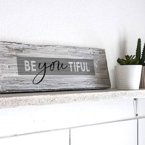 Könnte beinhalten: Ein weiß gestrichenes Holzschild mit dem Text "BE you TIFUL" in schwarzen Buchstaben. Das Schild steht auf einem weißen Regal mit zwei Topfpflanzen.