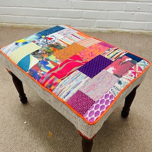 May include: A rectangular footstool featuring a patchwork top composed of various colourful fabric squares in shades of purple, orange, blue, and pink. The top is bordered by bright orange piping, and the base is supported by four dark brown wooden legs.