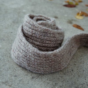 May include: A close-up of a light brown knitted scarf rolled up into a spiral shape. The scarf is lying on a gray concrete surface.