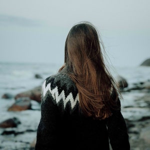 Op de afbeelding: Een vrouw met lang bruin haar staat op een rotsachtig strand, gekleed in een zwart-witte trui met patroon. De oceaan is op de achtergrond.