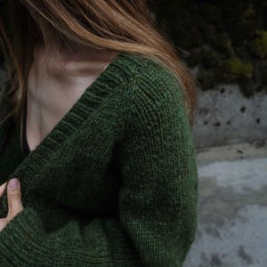Puede incluir: Un cárdigan de punto verde oscuro con cuello en V. El suéter está hecho de un hilo grueso y texturizado y tiene puños y dobladillo acanalados. El cárdigan es de color liso y parece hecho a mano.