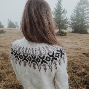 Puede incluir: Una mujer lleva un suéter de punto blanco con un patrón geométrico marrón y negro en el hombro. El suéter está hecho de un hilo suave y esponjoso. La mujer está de pie en un campo con árboles en el fondo.