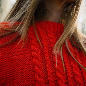 May include: A close-up of a red cable knit sweater. The sweater is made of a soft, chunky yarn and has a classic cable knit pattern.