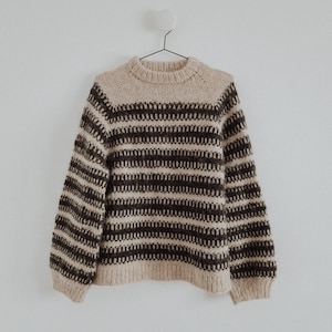 Op de afbeelding: Een gestreepte trui in beige en bruin met een ronde hals en lange mouwen. De trui hangt aan een witte hanger tegen een witte muur.