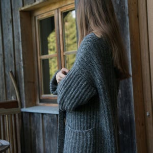 Könnte beinhalten: Ein langer, grauer Strickcardigan mit Rippendetails und zwei Vordertaschen. Der Cardigan wird von einer Person getragen, die sich in der Nähe eines Holzfensters und einer Holztür befindet. Die Gesamtästhetik ist gemütlich und rustikal.