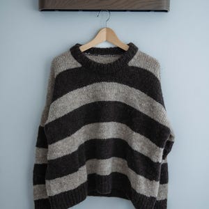 Peut inclure: Un pull en tricot épais à larges rayures horizontales marron foncé et marron clair est suspendu à un cintre en bois. Le pull a un col rond et des manches longues. Fond bleu clair avec une étagère en bois.