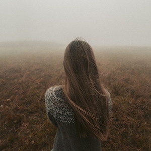 Puede incluir: Una persona con el pelo largo y castaño lleva un jersey de punto gris y se encuentra en un campo de hierba alta con niebla de fondo.