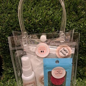 May include: A transparent tote bag containing beauty products. Inside are a white spray bottle, a bag of white powder, and a small blue pouch with two round, coloured items. The bag has a white button closure and clear handles.