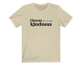Puede incluir: Una camiseta beige con la frase "Choose kindness" en negro, con una serie de puntos de colores encima de la palabra "kindness". La camiseta tiene mangas cortas y cuello redondo.