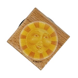 May include: A wooden square stamp with a yellow sun face with orange rays. The sun has a smiling face with closed eyes. The stamp is labeled "ME-E-INC."