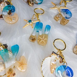 May include: A collection of beach-themed keychains with gold-colored hardware. The keychains feature resin designs with seashells, starfish, and sand, in shades of blue, beige, and clear. Some keychains are shaped like letters.