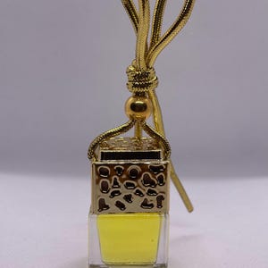 May include: A gold car air freshener with a yellow liquid inside. The air freshener has a gold chain with a gold bead at the top.