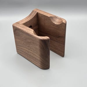 May include: A handcrafted wooden phone stand with a natural wood grain finish. The stand features a curved design with a slot to hold a phone and a small hole. The stand is made of solid wood.