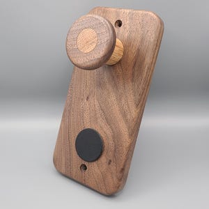 Może przedstawiać: Drewniany uchwyt ścienny z okrągłym pokrętłem i czarnym kółkiem w środku. Uchwyt wykonany jest z drewna orzechowego i ma naturalne wykończenie.