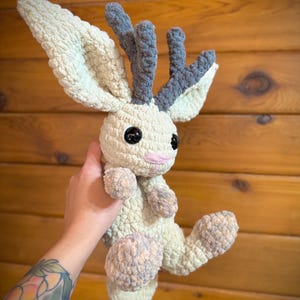 Pu&ograve; includere: Un peluche fatto a mano all'uncinetto a forma di creatura simile a un cervo. Il giocattolo presenta grandi orecchie e corna strutturate nei toni del crema e del grigio. Ha grandi occhi neri, una bocca rosa e arti morbidi e arrotondati.