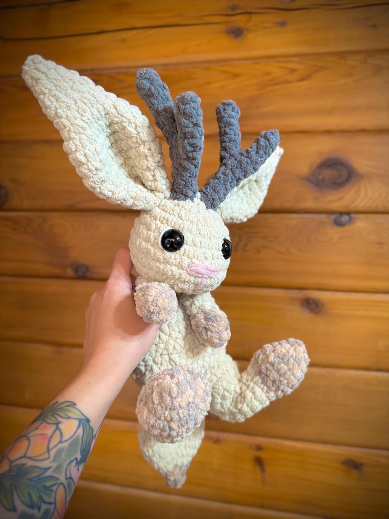 Pu&ograve; includere: Un peluche fatto a mano all'uncinetto a forma di creatura simile a un cervo. Il giocattolo presenta grandi orecchie e corna strutturate nei toni del crema e del grigio. Ha grandi occhi neri, una bocca rosa e arti morbidi e arrotondati.