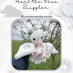 Könnte beinhalten: Eine weiße Häkeltaube, "Pearl the Dove Snuggler", mit rosa Füßen und Schnabel sowie blauen Augen. Das Bild enthält den Text "PDF PATTERN ONLY" und den Instagram-Handle @cozymountaincrochet. Die Taube wird vor einem Hintergrund aus grünem Gras und Bäumen gezeigt.