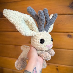 Pu&ograve; includere: Un peluche fatto a mano all'uncinetto a forma di creatura simile a un cervo. Il giocattolo presenta grandi orecchie flosce, corna grigie e occhi neri a bottone. Il corpo &egrave; color crema con accenti grigi e rosa.